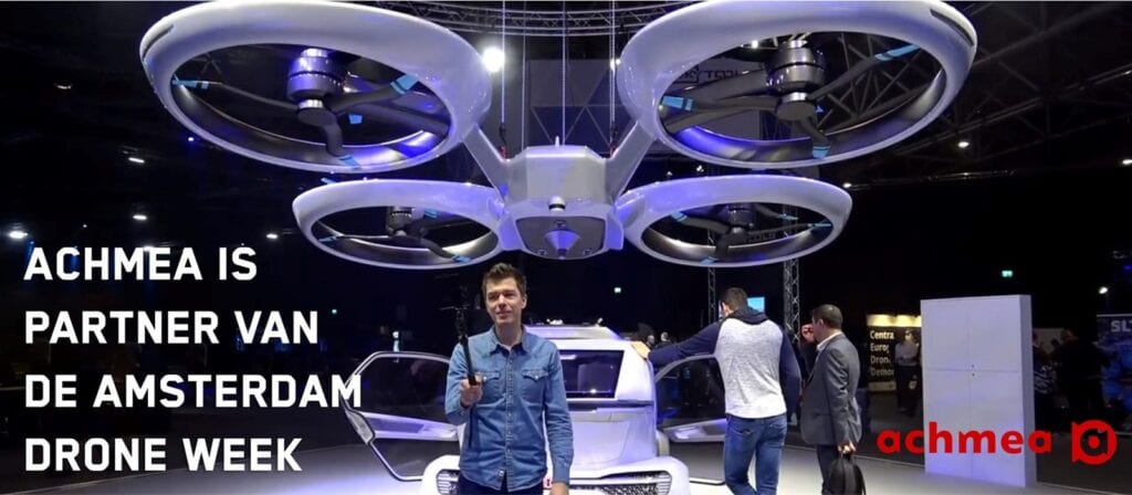 A man stands in front of a large drone and futuristic vehicle at an event. Text reads Achmea is partner van de Amsterdam Drone Week. The Achmea logo is visible in the lower right corner.