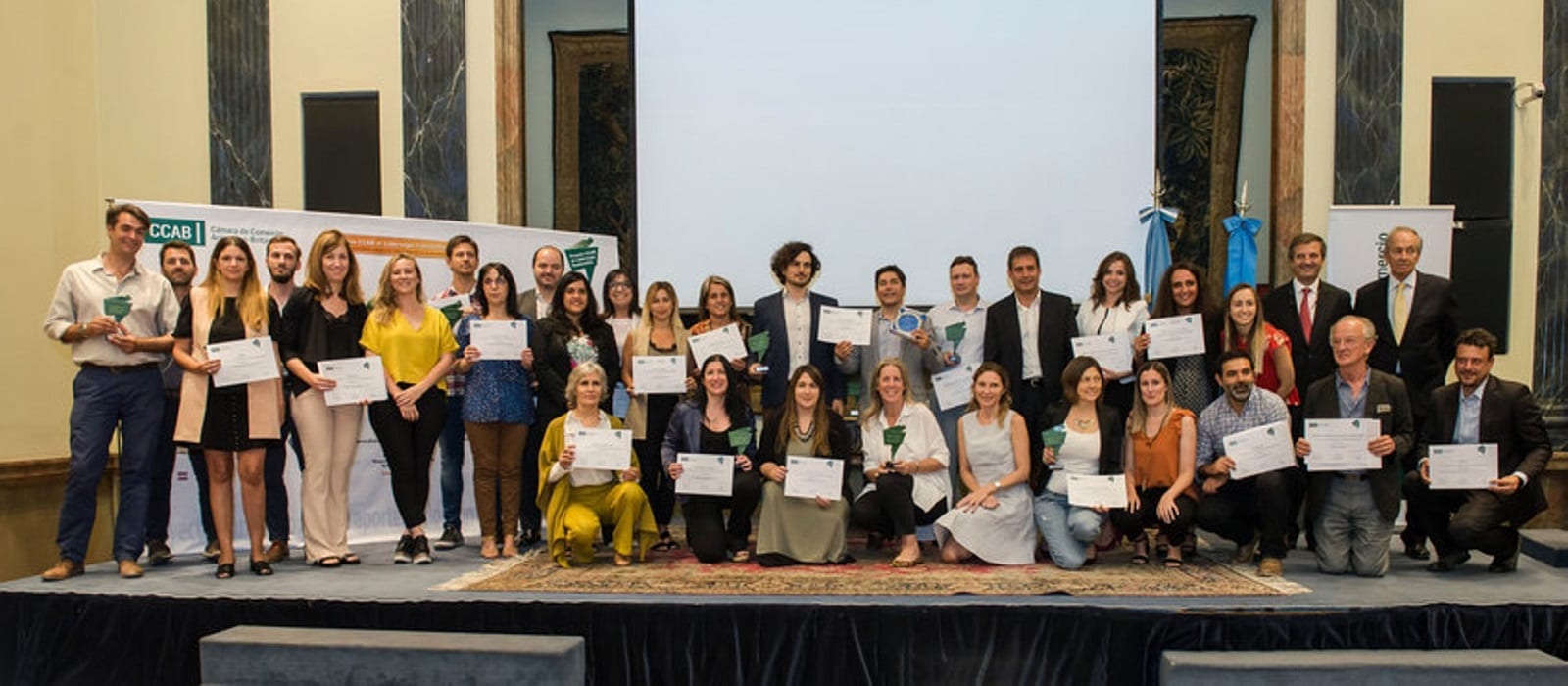 A group of people pose on stage holding certificates and trophies, standing and kneeling in rows, in front of a blank projector screen and banners at an indoor awards or graduation event.