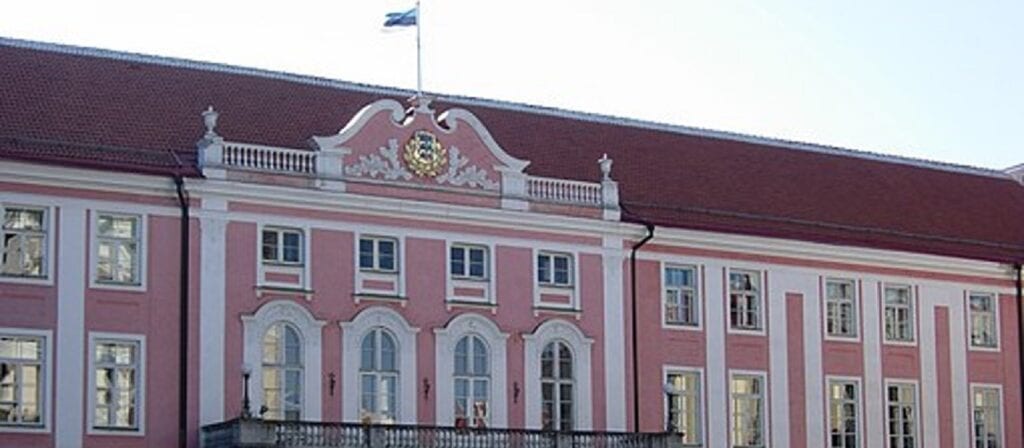 A historic pink and white building with arched windows, a decorative crest above the entrance, and a flag flying on the roof against a clear sky.