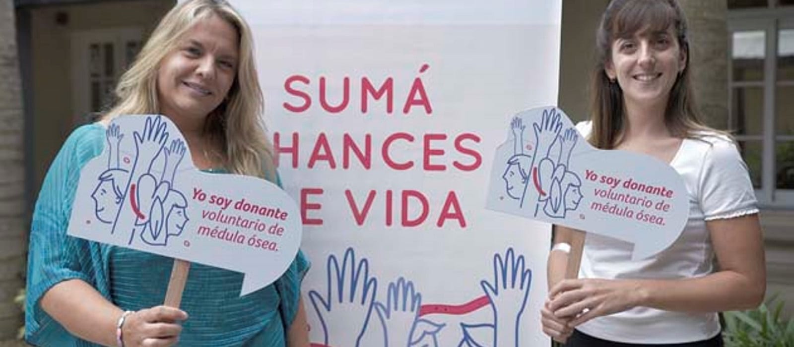 Two women smile and hold signs that read Yo soy donante voluntario de médula ósea in front of a banner with the words Sumá Chances de Vida and illustrations of raised hands.