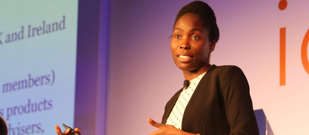 A woman holding a remote control speaks on stage in front of a projected screen displaying text. She is wearing a black jacket and white blouse, and appears to be giving a presentation or lecture.