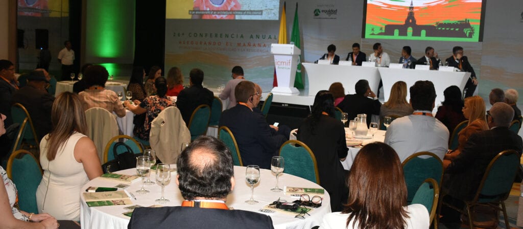 People sit at round tables facing a panel of speakers on a stage at a conference. A large screen and banners are visible behind the speakers, and the setting appears formal with business attire and conference materials.