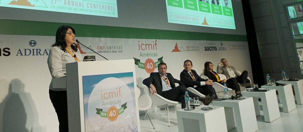 A woman stands at a podium speaking to an audience at the ICMIF Americas Conference. Four seated panelists listen on stage, with a large conference banner displayed behind them.