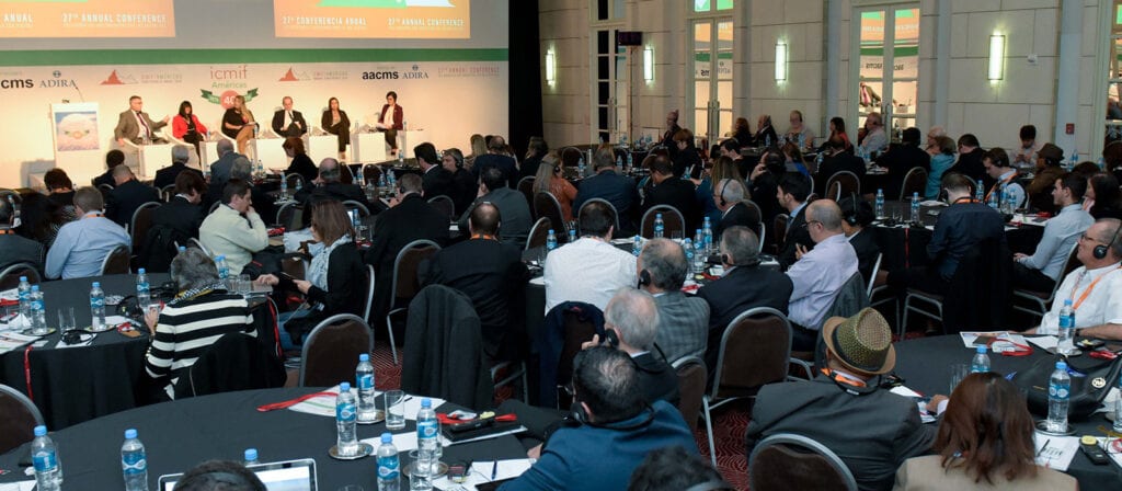 A large conference room filled with attendees seated at round tables, facing a stage where six panelists are speaking. Bottles of water and conference materials are on the tables.