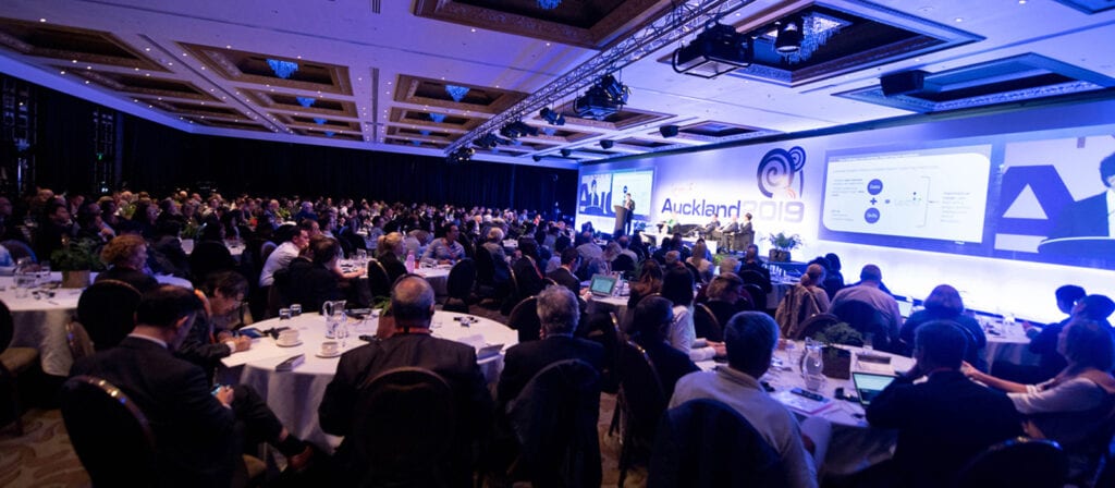 A large audience sits at round tables in a conference hall, watching a panel discussion on stage with presentations projected on screens at an event labeled Auckland 2019.