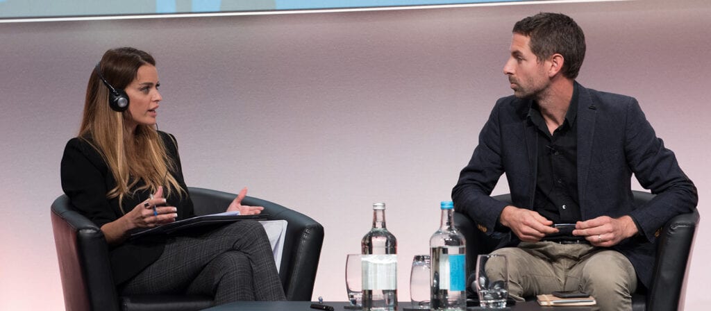 Two people sit in black chairs on a stage, engaged in conversation. The woman on the left wears headphones and holds papers, while the man on the right listens. Bottles and glasses are on the table between them.