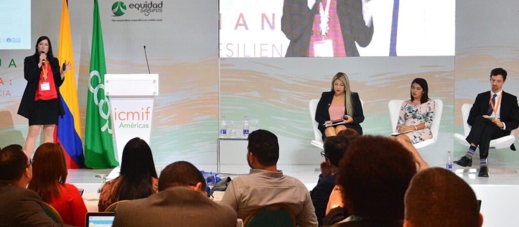 A woman stands and speaks at a podium on stage, while three seated panelists listen. An audience faces them, and flags and a conference backdrop are visible behind the speakers.