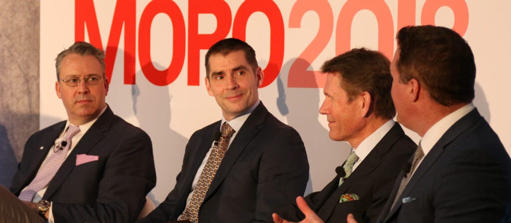 Four men in business suits sit and talk on a panel in front of a large red and white MORO 2018 sign. They appear engaged in discussion, with one man speaking as the others listen attentively.