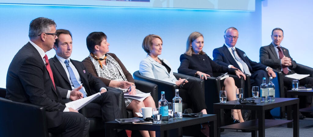 Six professionally dressed individuals sit on a stage panel, engaging in discussion. They are seated in black chairs with microphones and water bottles on tables in front of them. The background is light blue.
