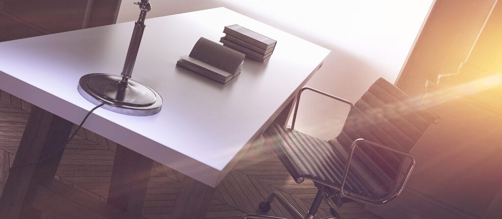A modern office desk with a metal lamp, three stacked books, an open notebook, and a black swivel chair. Sunlight streams through a window, casting a warm glow and shadows across the space.