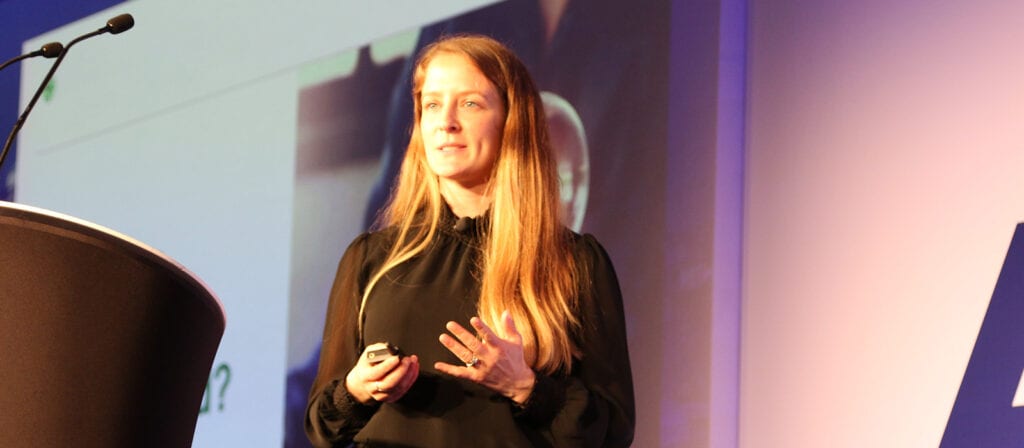 A woman with long blonde hair stands on stage holding a remote, giving a presentation. She is wearing a black blouse, and a microphone and podium are visible. A projected slide is blurred in the background.
