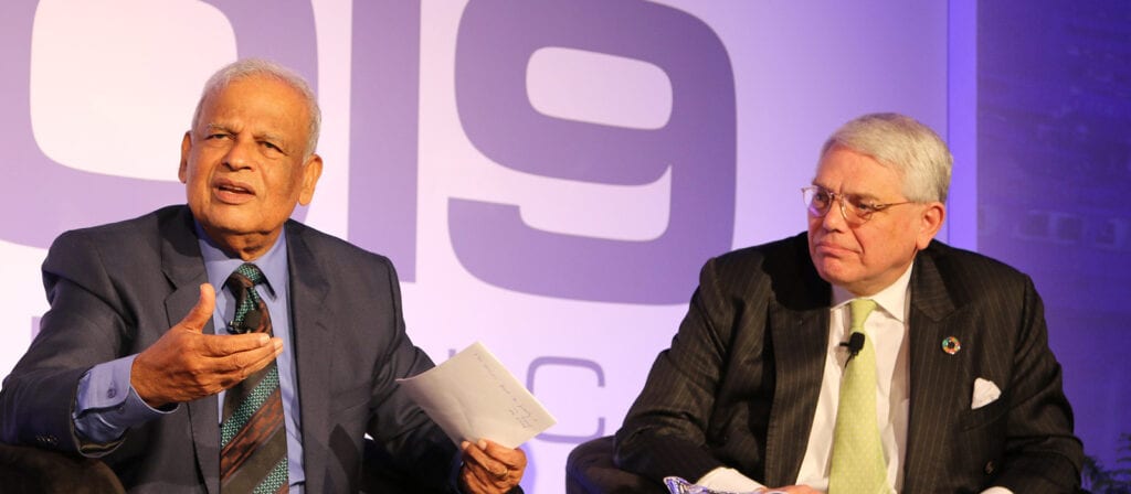 Two older men in suits sit on a stage in front of a large 2019 sign. One man gestures while speaking and holding papers, and the other listens attentively, hands folded in his lap.