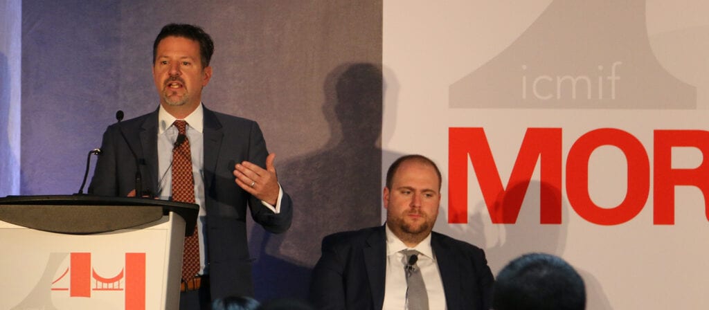Two men in suits are at a conference; one stands and speaks at a podium, gesturing with his hand, while the other sits and listens. A large sign behind them displays icmif and MOR.