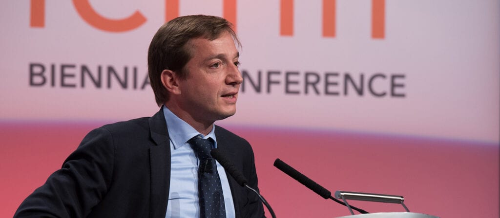 A man in a suit and tie speaks at a podium during a conference, with a microphone in front of him and a screen behind displaying the words “BIENNIAL CONFERENCE.”.