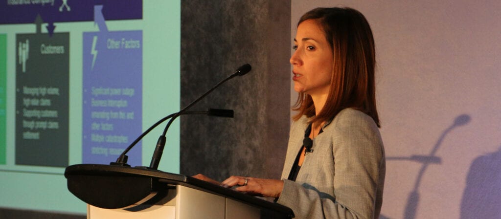 A woman in a gray blazer stands at a podium speaking into a microphone, with a projected presentation slide featuring text and graphics visible in the background.