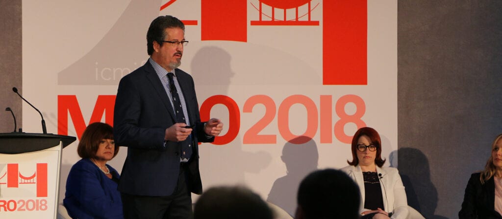 A man in a suit speaks on stage at a conference, with three women seated behind him. A large red and white sign in the background reads MO2018.