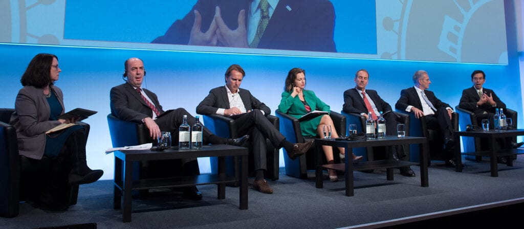 Six people in business attire sit on stage in armchairs, participating in a panel discussion. There are water bottles and glasses on tables in front of them, and a large screen behind them displays part of an image.