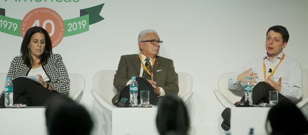 Three people sit on a panel at a conference, two men and one woman, with microphones and water bottles in front of them. One man is speaking while the others listen. A banner behind them reads 40 years 1979–2019.