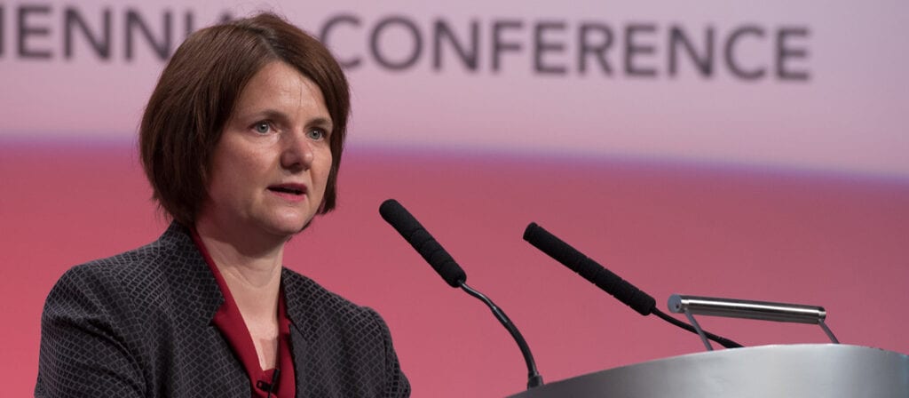 A woman with short brown hair and a maroon top speaks at a podium with two microphones in front of a blurred background displaying the words CONFERENCE.