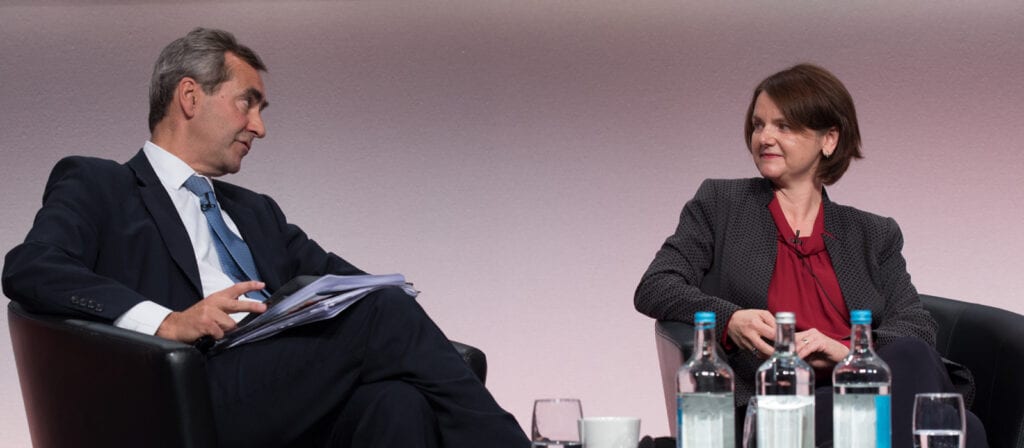 A man in a suit and a woman in business attire sit in black armchairs on a stage, engaged in conversation. There are water bottles and glasses on a table in front of them.