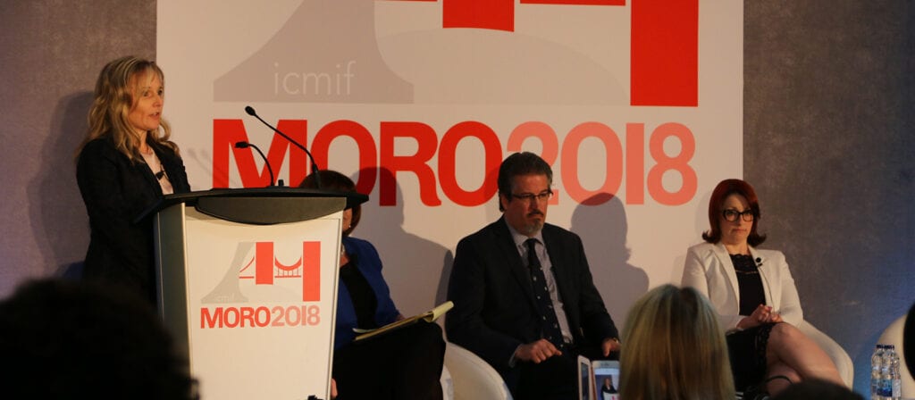 Four people sit on a stage at a conference. One woman is speaking at a podium labeled MORO2018. Behind them is a large event banner with the same text and the ICMIF logo. The audience is visible in the foreground.
