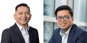 Two men in business attire, both wearing glasses and navy suits with white shirts, smiling. The man on the left has short hair and no glasses; the man on the right has dark hair and glasses, seated by a window.