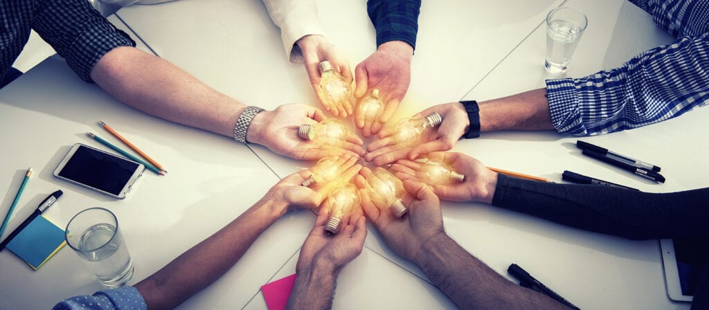 A group of people seated around a table extend their hands to the center, each holding a glowing light bulb, symbolizing teamwork and collaboration. Various office items and glasses of water are on the table.