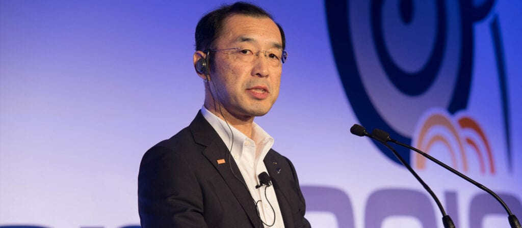 A man in a suit and glasses speaks at a podium with microphones. He is wearing an earpiece and stands in front of a blue and white background with circular designs and partial text.