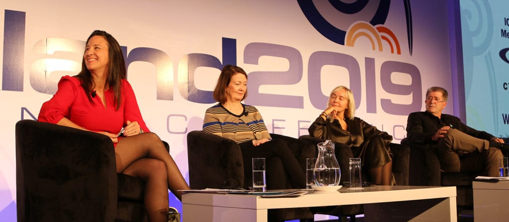 Four people sit on stage in armchairs during a panel discussion at a conference, with a large 2019 and part of the word Conference visible on the backdrop behind them. A table with water glasses and papers is in front of them.