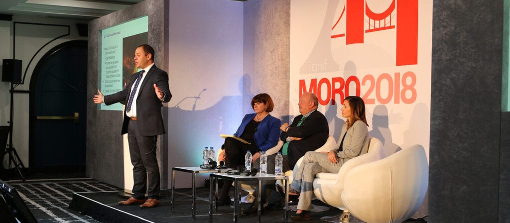 A man in a suit speaks onstage during a panel discussion at MORO 2018, while three seated panelists listen. A presentation slide is projected on the wall beside him.