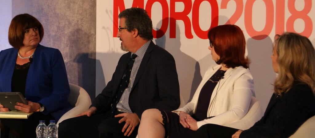 Four people in formal attire sit on a stage panel, engaged in conversation. A woman on the left holds a tablet, while the others listen. The background has large red text displaying MORO2018.