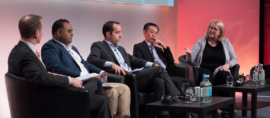 Five people in business attire sit on a panel stage, engaged in discussion. One woman gestures while speaking, and the four men listen attentively. Bottled water and glasses are on the table in front of them.