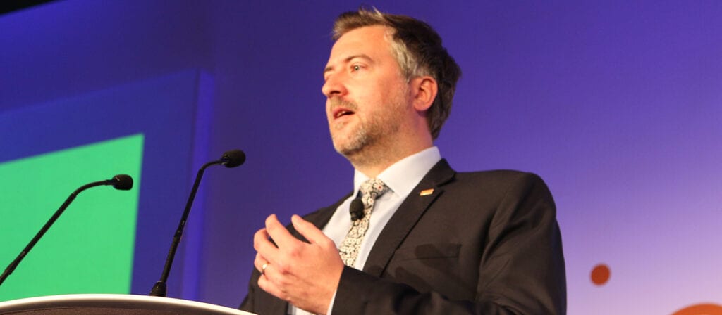A man in a suit and tie speaks at a podium with two microphones against a purple background with a green rectangular screen.