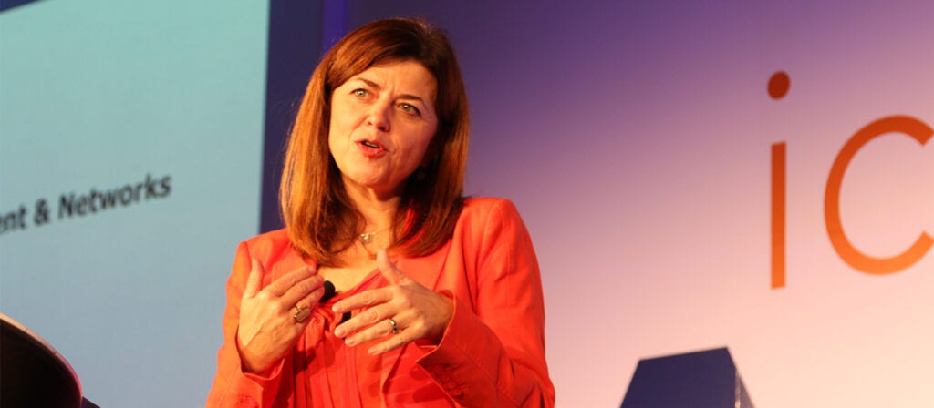 A woman with brown hair wearing an orange blazer speaks on stage, gesturing with her hands. A blurred presentation screen and partial text are visible in the background.