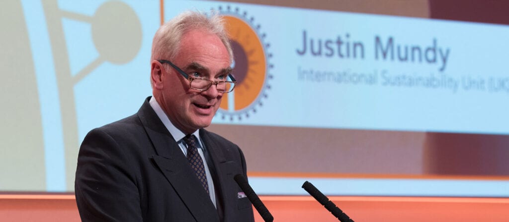 A man in a suit and glasses speaks at a podium, with a screen behind him displaying the name Justin Mundy and International Sustainability Unit (ISU).