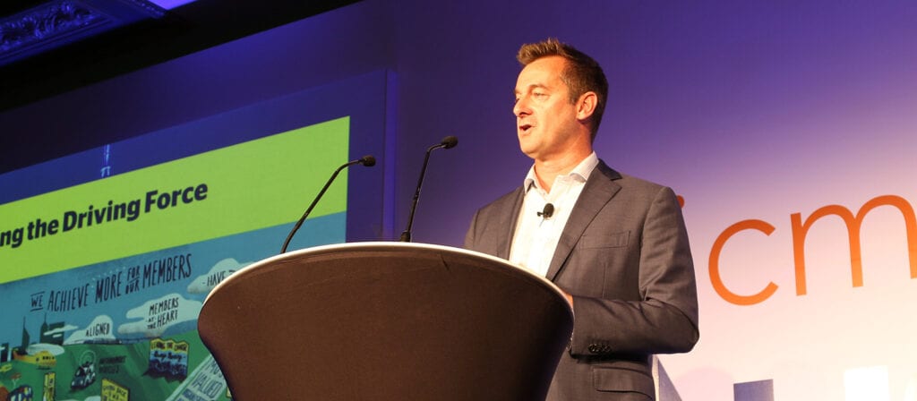 A man in a suit stands at a podium with two microphones, speaking at a conference. A colorful presentation slide and part of the letters cm are visible in the background.