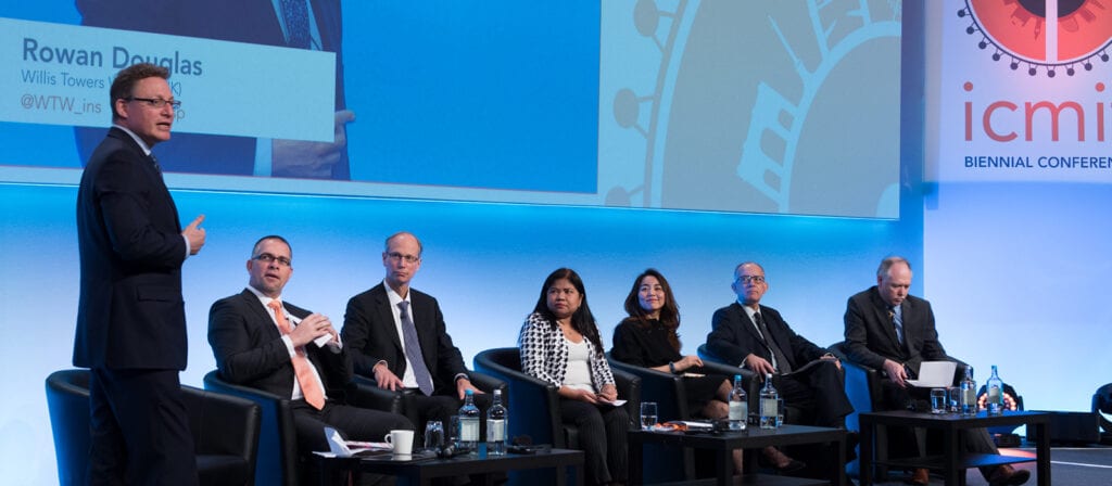 A man stands and speaks onstage to an audience while six other panelists, both men and women, sit and listen. A large screen behind displays the name Rowan Douglas and conference branding.