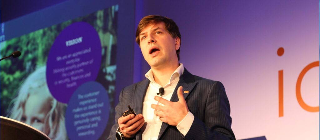 A man in a suit stands on stage giving a presentation, speaking into a headset microphone and holding a remote. A blurred presentation slide with text and images is visible in the background.