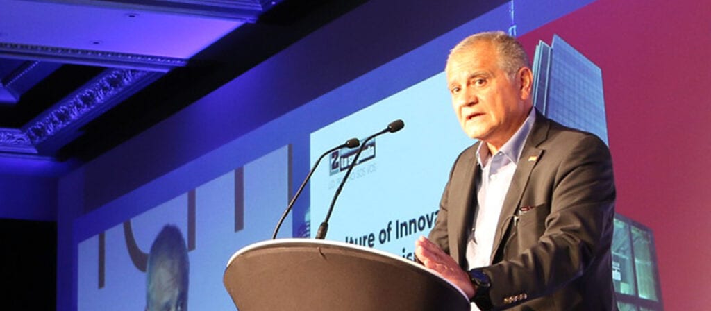 A man in a suit speaks at a podium with microphones at a conference. A presentation slide with text and a modern building is projected behind him. The event appears to be held in a formal indoor setting.