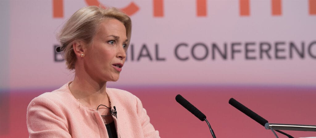 A woman with short blonde hair, wearing a light pink blazer, speaks at a podium with two microphones in front of a blurred conference backdrop.