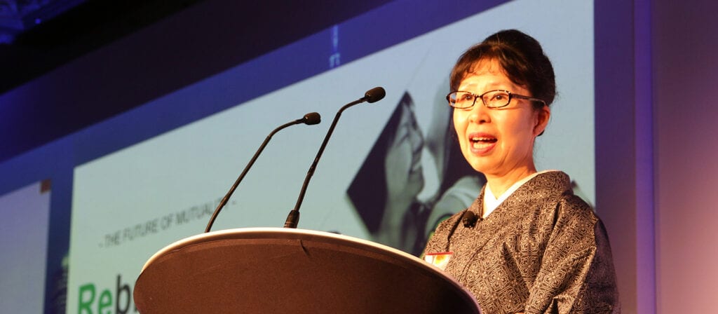 A woman wearing glasses and a patterned garment speaks at a podium with two microphones, in front of a projected presentation slide on a large screen.