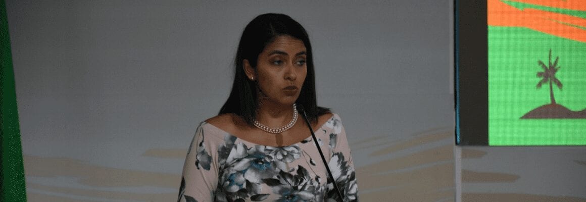A woman with long dark hair, wearing a floral dress and a pearl necklace, stands at a podium speaking. A green and orange screen with a palm tree graphic is partially visible in the background.