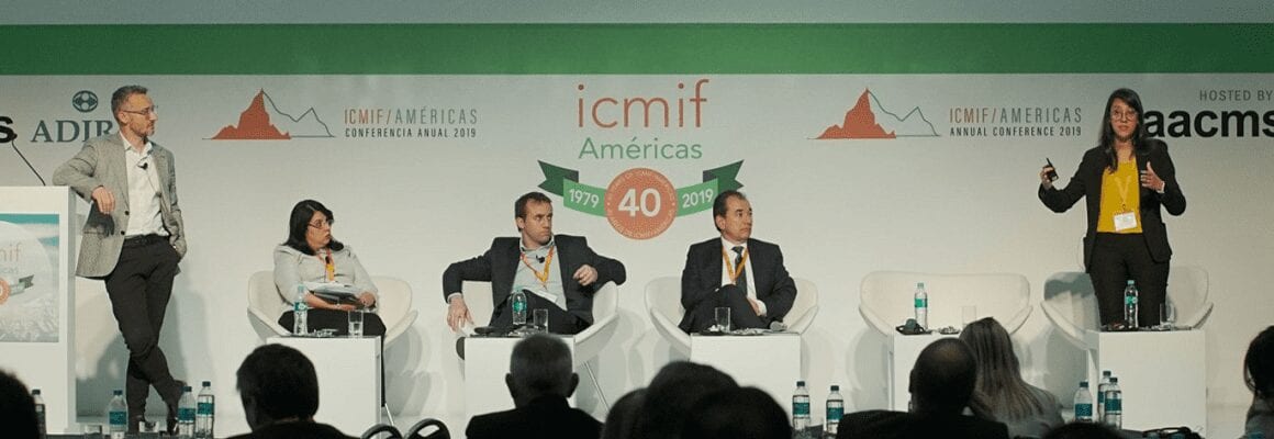 Five panelists sit on stage at the ICMIF Americas conference; one woman stands and speaks at a podium while the others listen. A presentation banner and audience members are visible.