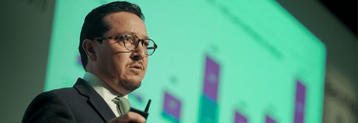 A man in a suit and glasses speaks while holding a remote, standing in front of a projected bar graph with purple and green sections. The background is blurred, focusing on the speaker.