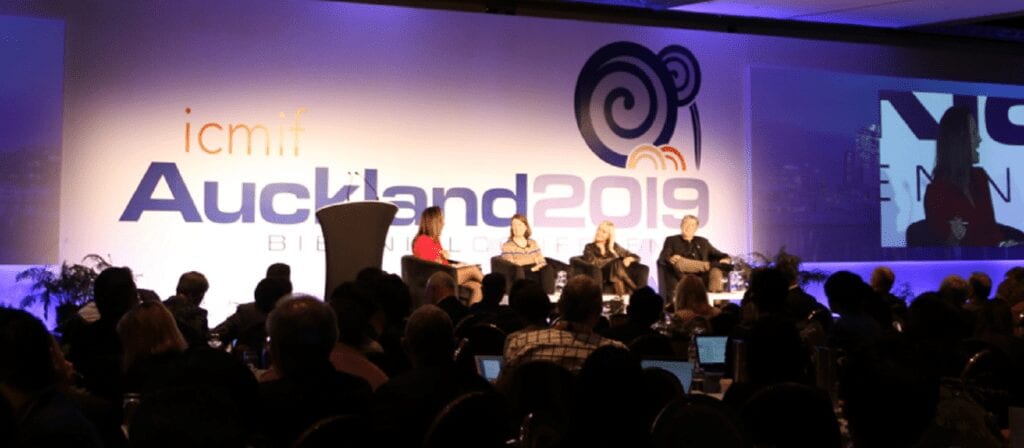 A panel of four people are seated onstage at the ICMIF Auckland 2019 conference, speaking to an audience in a dimly lit room with a large event banner and projected screen behind them.
