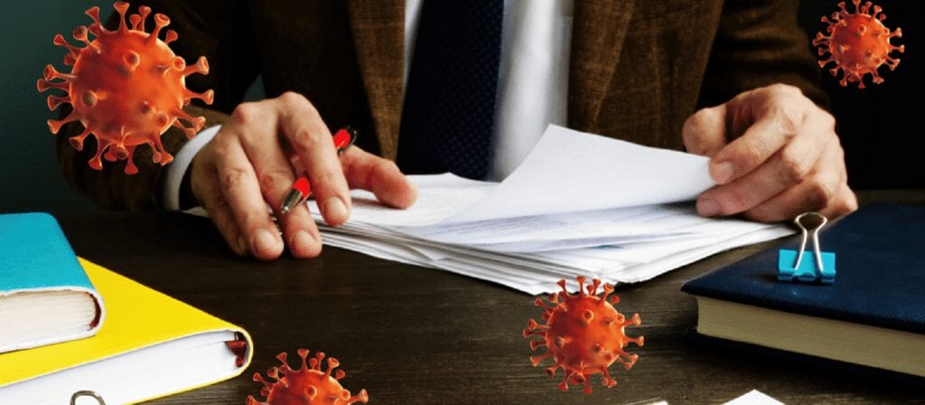 A person in a suit reviews paperwork at a desk with books and folders, while illustrated virus particles float in the foreground, symbolizing the impact of COVID-19 on office work.