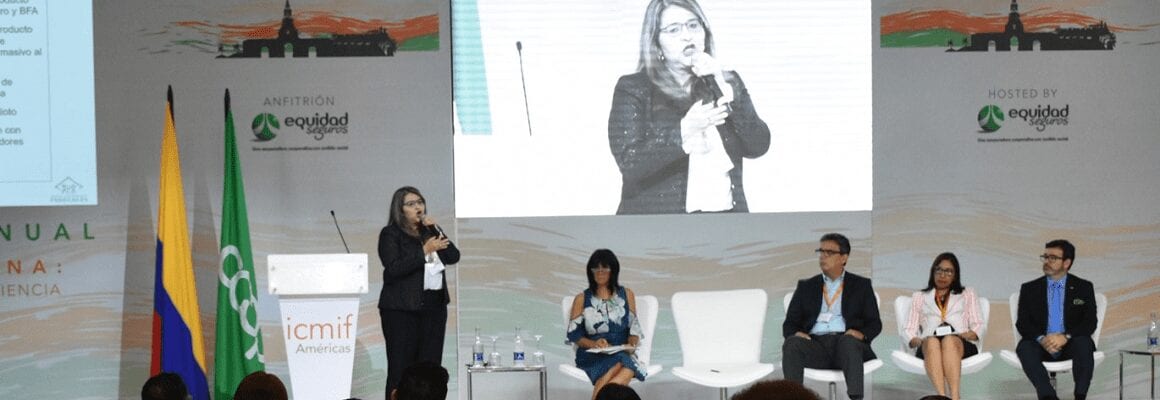 A woman speaks into a microphone on stage beside four seated panelists at a conference. Behind them are large screens showing her image, flags, and event banners with logos and text in Spanish.