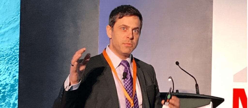 A man in a suit and tie speaks at a podium during a conference, gesturing with one hand and holding his glasses in the other. He has a serious expression and is wearing an event lanyard.