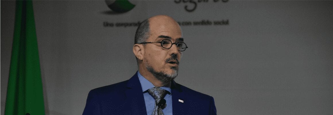 A man with glasses and a beard, wearing a blue suit and tie, speaks in front of a backdrop with partially visible text and a green flag.