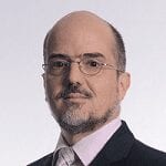 A middle-aged man with glasses, a trimmed beard, and a bald head, wearing a dark suit, light shirt, and tie, posing against a plain light background.